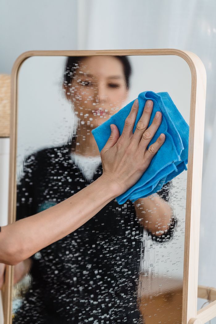 hero-img-01 Close-up view of a person cleaning a mirror with a blue cloth, with hands and reflection visible.